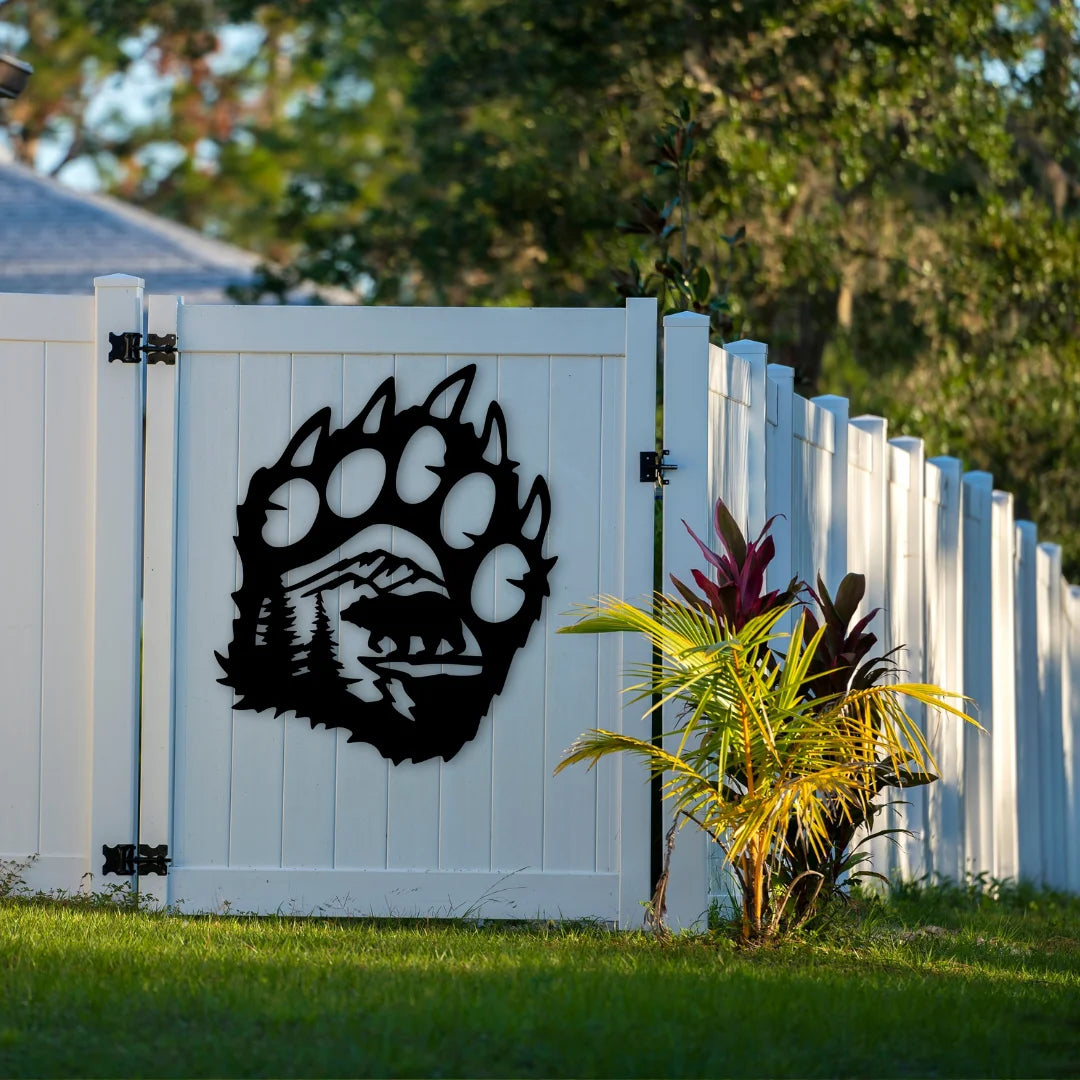 A black bear paw shaped outdoor metal decor featuring a landscape with mountains and trees stands out on a white fence, adding wild charm to a garden entry.