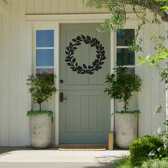 A minimalist black metal outdoor decoration in the shape of a leafy wreath is mounted on a pale green door, offering timeless elegance to a cozy front porch.