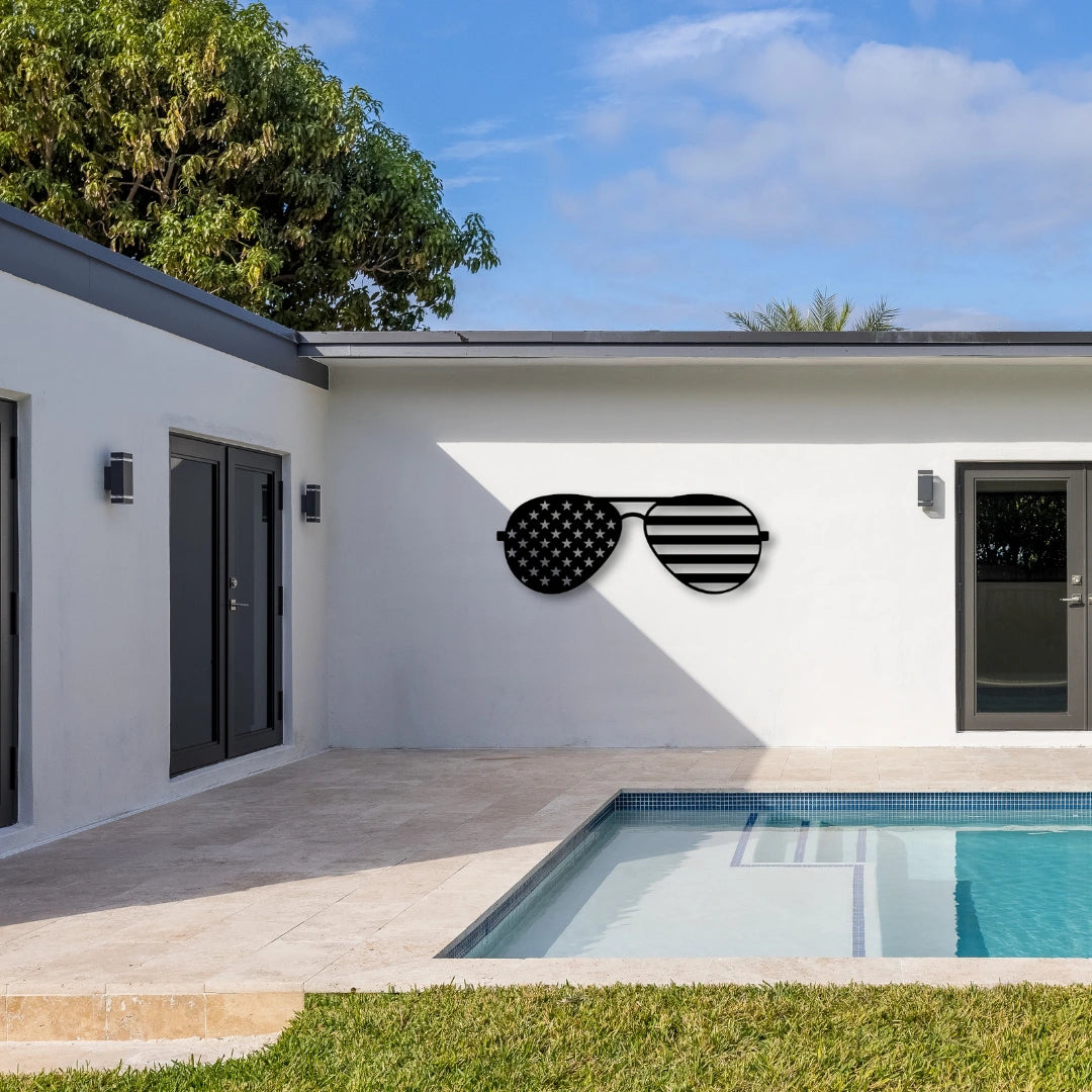 Patriotic metal wall art shaped like aviator sunglasses with stars and stripes decorates a white poolside wall, merging outdoor cool with patriotic designs.