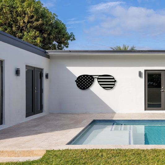 Patriotic metal wall art shaped like aviator sunglasses with stars and stripes decorates a white poolside wall, merging outdoor cool with patriotic designs.