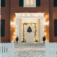 A festive metal christmas sign in a tree shape with monogram hangs on a snowy front door, surrounded by glowing lights and pine trim — a striking metal sign for holiday homes.
