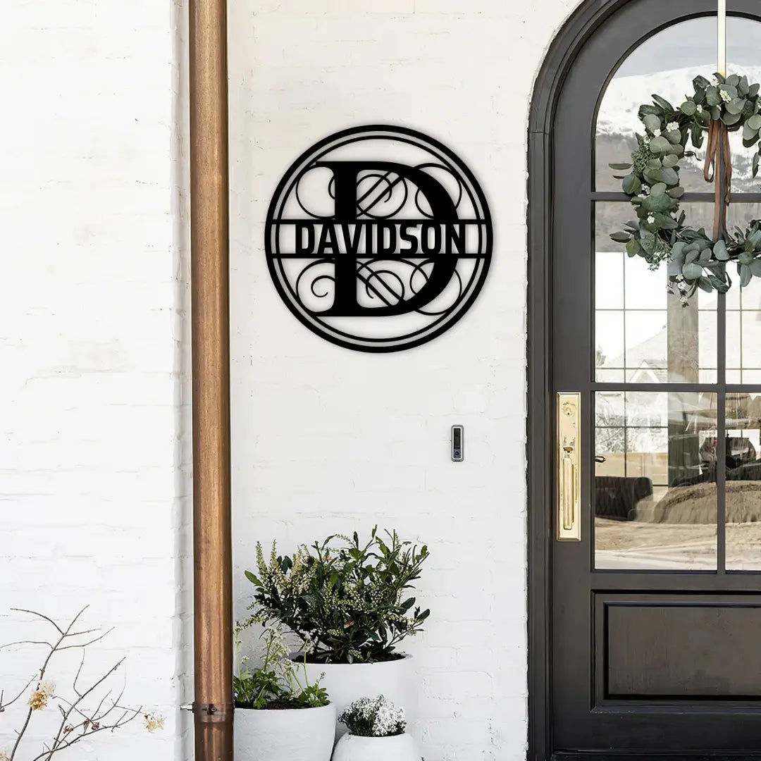 A black monogram signs circle with an ornate initial and surname sits beside a modern entryway, pairing beautifully with custom planters and personalized metal signs style.