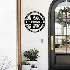 A black monogram signs circle with an ornate initial and surname sits beside a modern entryway, pairing beautifully with custom planters and personalized metal signs style.