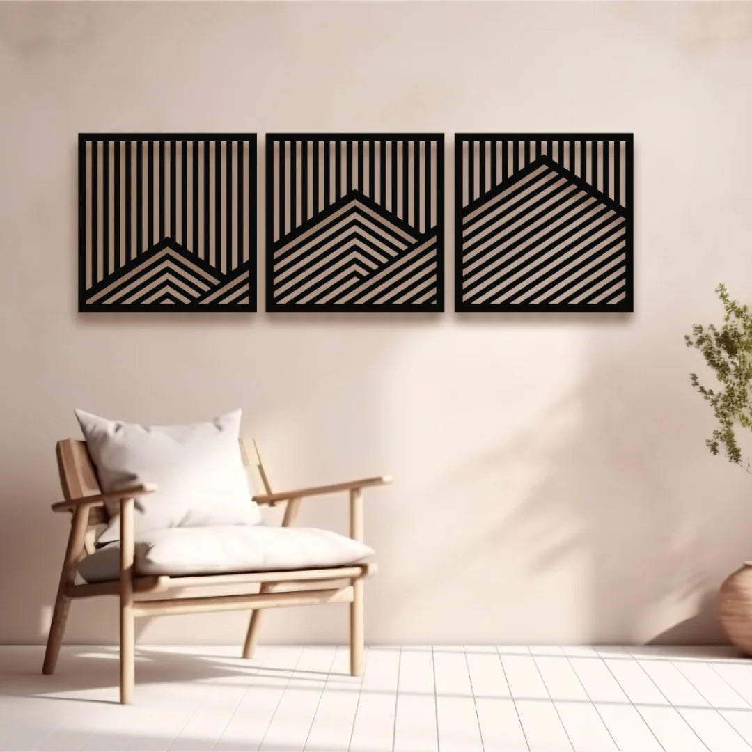 A set of three decorative metal panel artworks with diagonal and vertical lines forming abstract mountain shapes, mounted on a soft beige wall above a minimal wooden armchair.