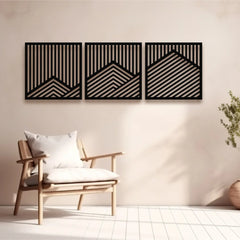 A set of three decorative metal panel artworks with diagonal and vertical lines forming abstract mountain shapes, mounted on a soft beige wall above a minimal wooden armchair.