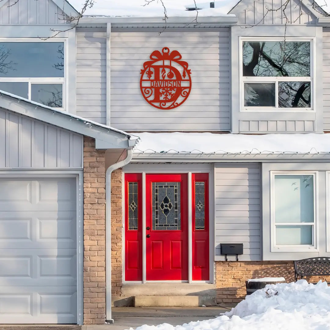 A festive red front door stands out on a snowy porch, with a round custom metal yard sign above, featuring a personalized house address sign and ribbon-inspired design perfect for holiday decor.
