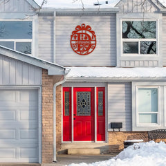 A festive red front door stands out on a snowy porch, with a round custom metal yard sign above, featuring a personalized house address sign and ribbon-inspired design perfect for holiday decor.