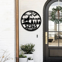 A round address sign for yard featuring a howling wolf, pine forest, and mountain silhouette, mounted beside a black front door on a white brick wall.