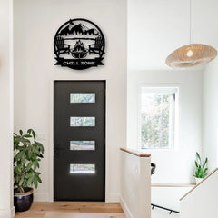 A round Chill Zone steel wall art featuring Adirondack chairs and a firepit, mounted above a contemporary black front door, creating a cozy, welcoming vibe.