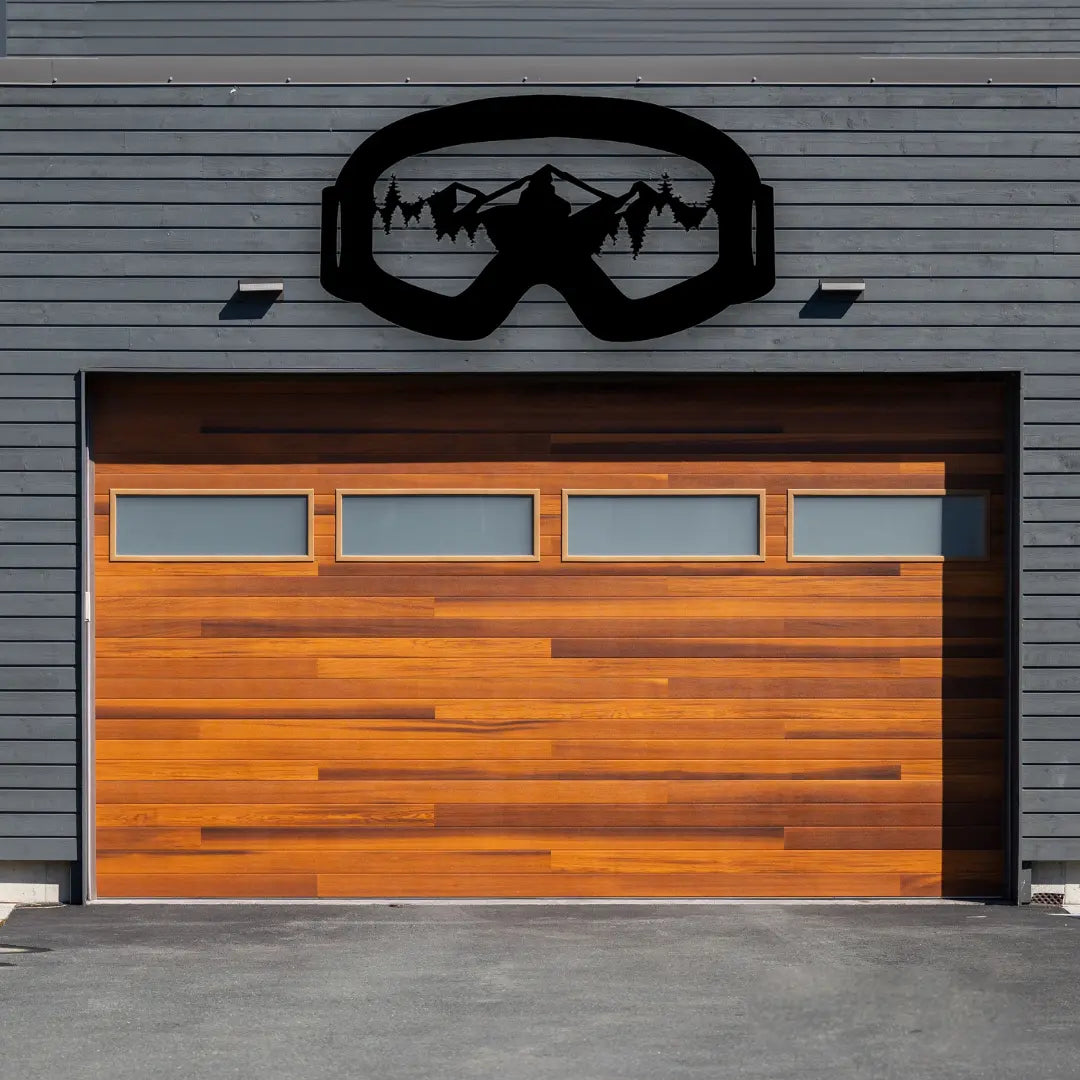 Above a wood-accent garage, this metal mountain wall art shaped like ski goggles reveals detailed peaks and forest lines, embodying bold mountain artwork for adventurers.