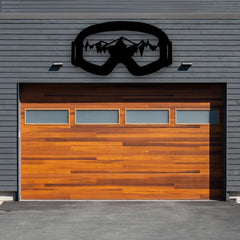 Above a wood-accent garage, this metal mountain wall art shaped like ski goggles reveals detailed peaks and forest lines, embodying bold mountain artwork for adventurers.