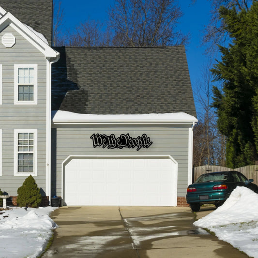 A bold script patriotic wall art reading “We the People” hangs beside a white brick doorway, representing national pride through metal outdoor signs for entry decor.