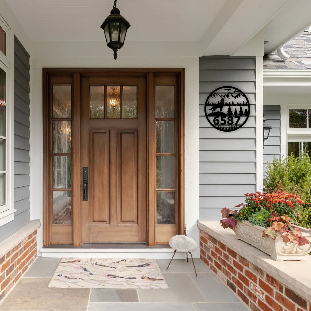 A round-shaped custom metal address sign with wildlife and mountain elements displays the house number in a rustic, nature-inspired style.
