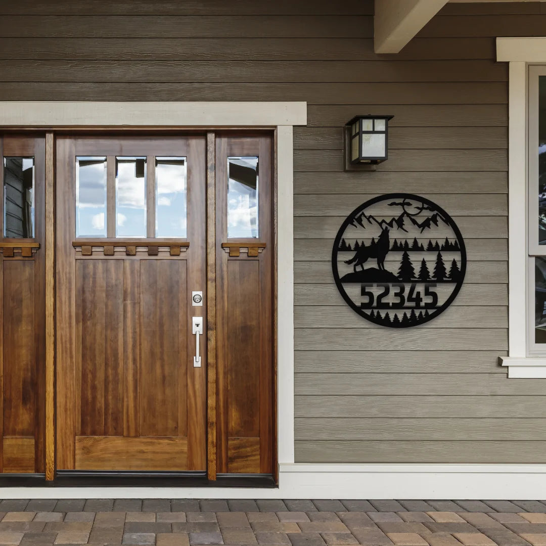 A round custom address sign for yard with howling wolf, trees, and mountains features a house number, mounted near a wooden entry door for a rustic outdoor look.
