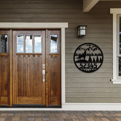 A round custom address sign for yard with howling wolf, trees, and mountains features a house number, mounted near a wooden entry door for a rustic outdoor look.
