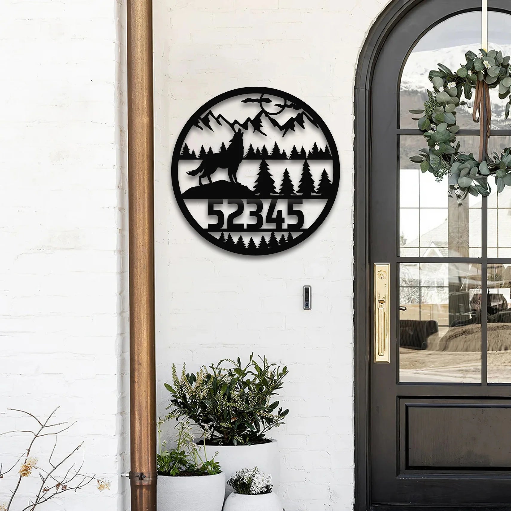 A round address sign for yard featuring a howling wolf, pine forest, and mountain silhouette, mounted beside a black front door on a white brick wall.

