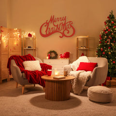 A red merry christmas sign decorates the wall above two cozy chairs in a warmly lit holiday room with a glowing Christmas tree and wrapped gifts.
